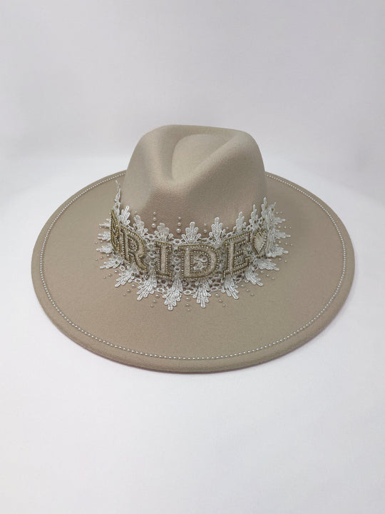 Beige fedora hat featuring interchangeable handmade lace bands with Velcro attachments. Includes three designs: “BRIDE” and “WIFEY” lettering embellished with crystals and pearls, and an elegant floral motif with lace, handmade  flowers and beaded details. A stylish 3-in-1 bridal accessory made in the UK. One size, wide brim fedora, luxury fedora, bride hat, wifey hat, floral hat, hen do
