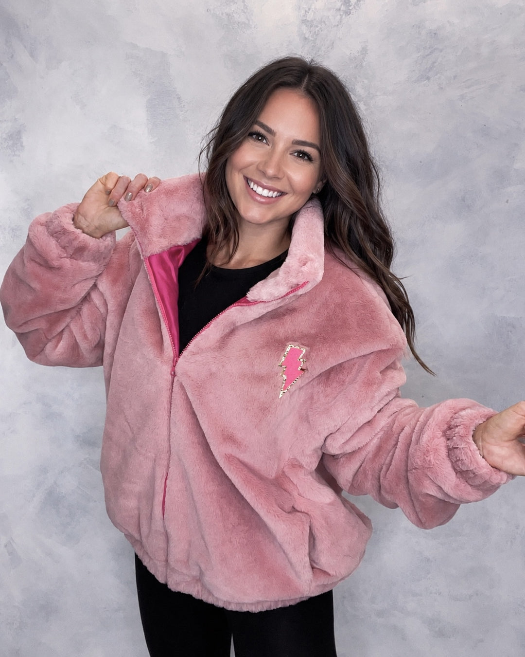 A person wearing a pink faux fur bomber jacket with 'VIBE' lettering embroidered on the back in glitter. Festival jacket, fur coat, winter coat