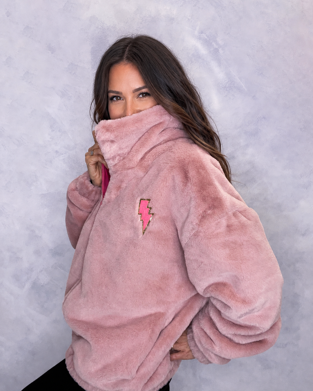 A woman wearing a plush, dusty pink faux fur jacket with a small pink lightning-bolt patch on the chest. festival jacket, winter coat