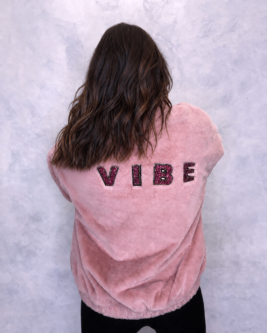 A person wearing a pink faux fur bomber jacket with 'VIBE' lettering embroidered on the back in glitter. Festival jacket, fur coat, winter coat